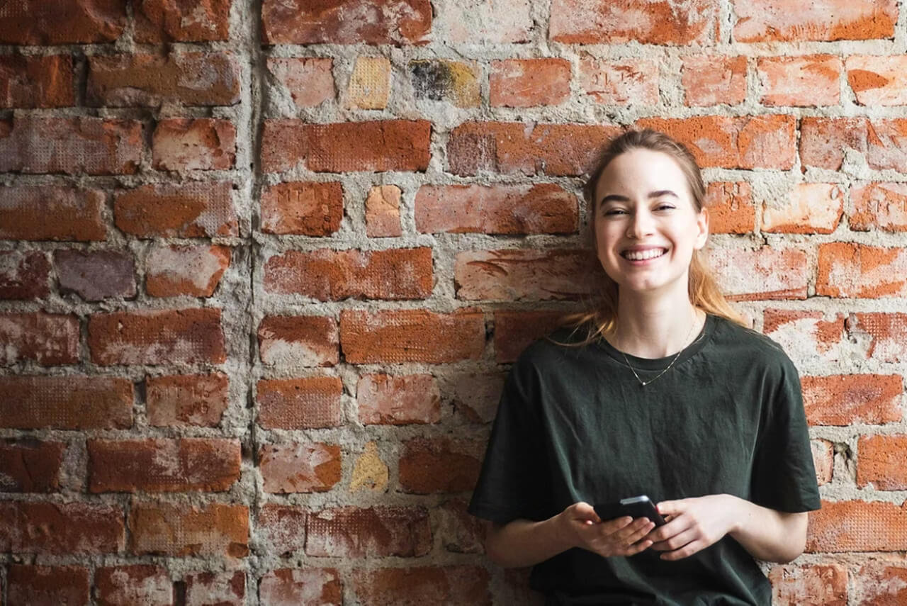 Young female with a phone stood against a brick wall