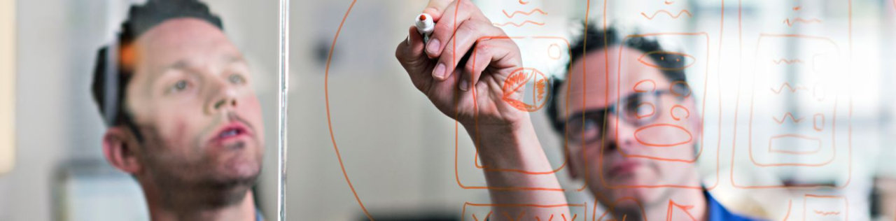 man drawing on glass board infront of colleauge